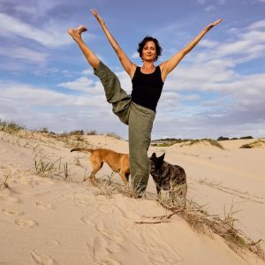Flexible woman lifting one leg out out to the side in a Yoga pose, with two dogs, standing on the sand.
